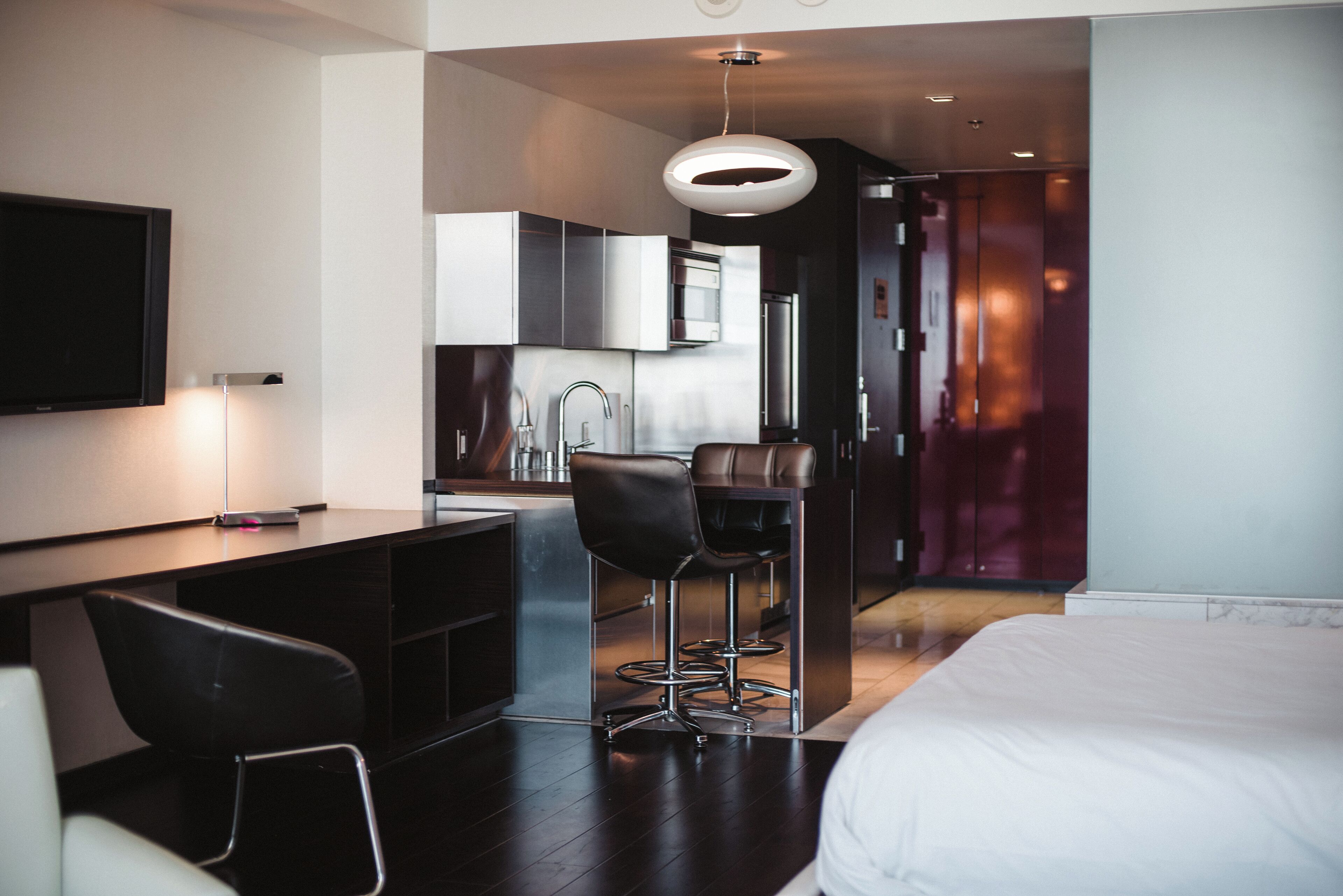 A modern hotel room with a large bed, a desk with a chair, a TV, and a kitchenette area.