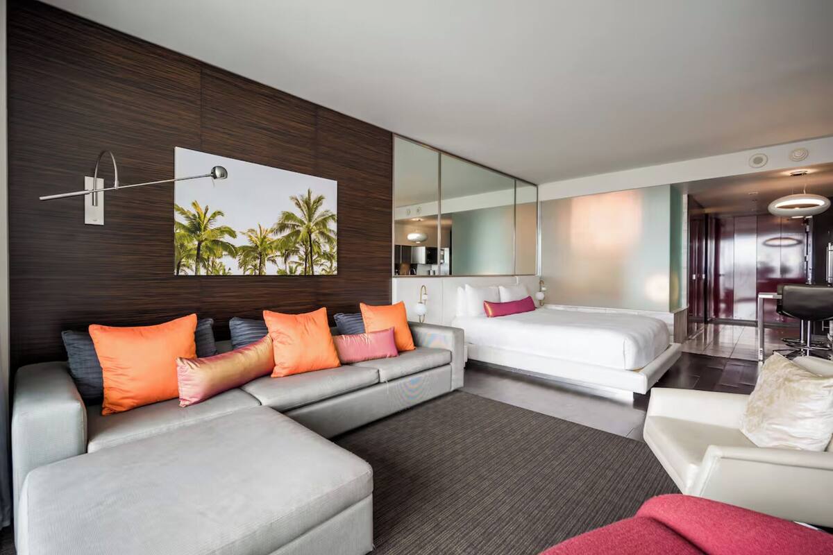 Une chambre d’hôtel moderne avec un canapé gris, un lit blanc et un grand miroir.