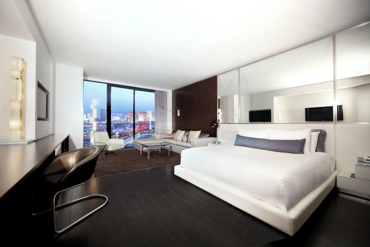 Une chambre d’hôtel moderne dotée d’un grand lit, d’un bureau avec une chaise, d’un canapé et offrant une vue sur la ville grâce à de grandes fenêtres.