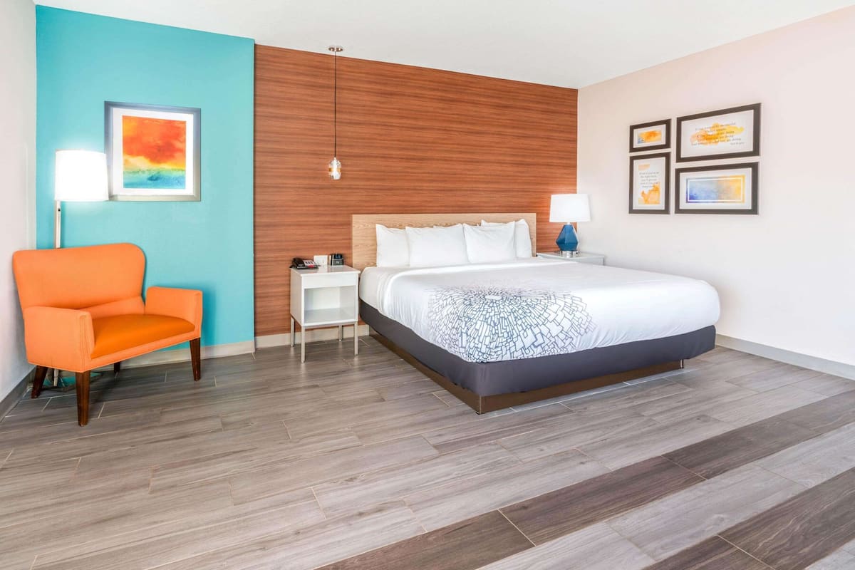 A modern hotel room with a large bed, a bright orange chair, a nightstand, and a wall with framed artwork.