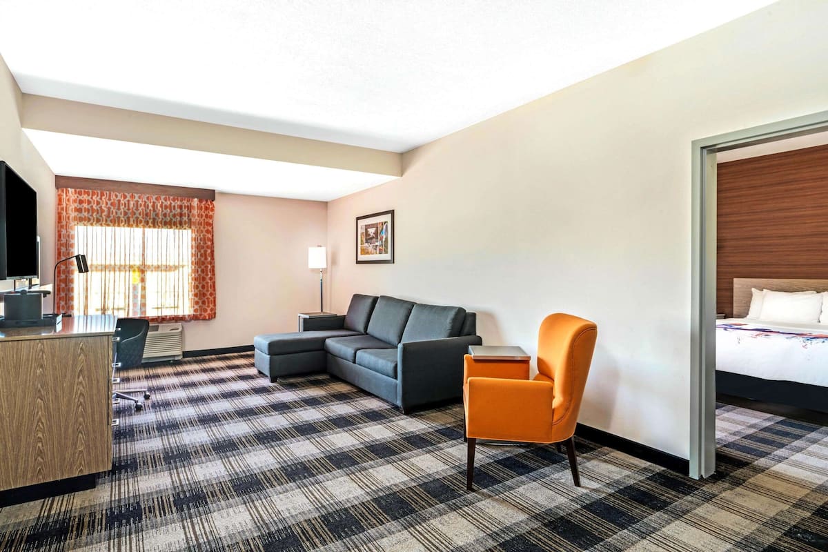 A hotel room with a check-in desk, a sofa, an orange armchair, a bed, and a large window with curtains.