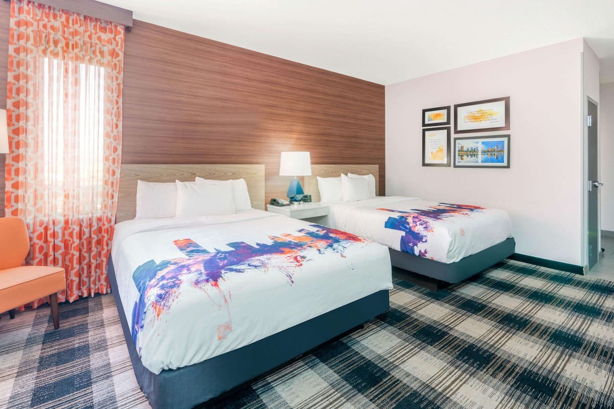 A hotel room with two beds, a nightstand, a lamp, a chair, and a plaid carpet.