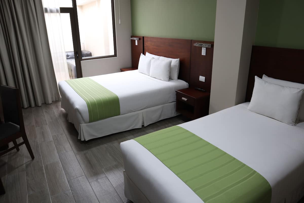 Habitación de hotel con dos camas, suelo de madera, pared verde y una ventana con cortinas.