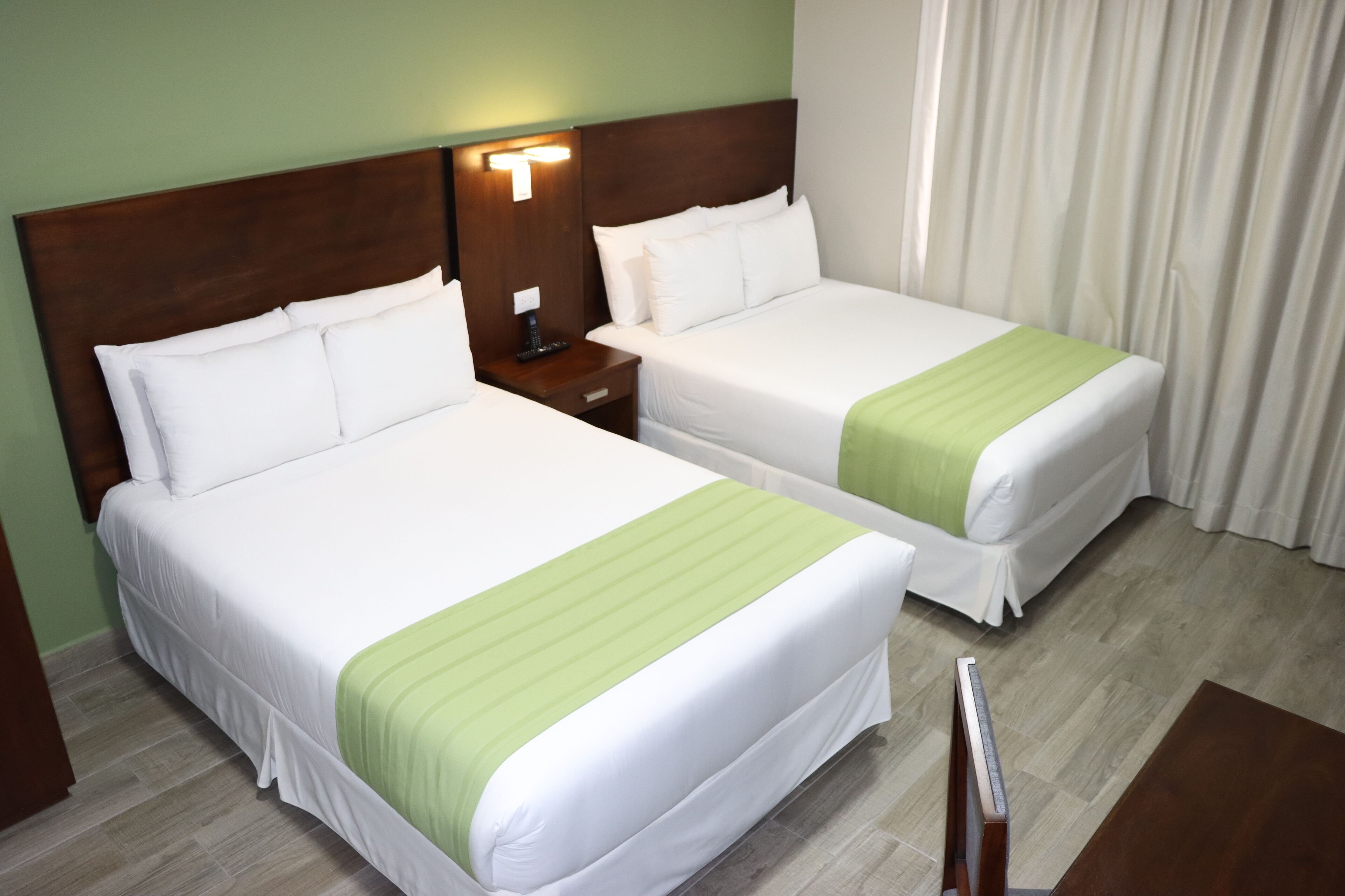 Deux lits simples avec du linge de lit blanc et des couvre-lits verts, une tête de lit en bois, une table de chevet avec une lampe et un plancher en bois.