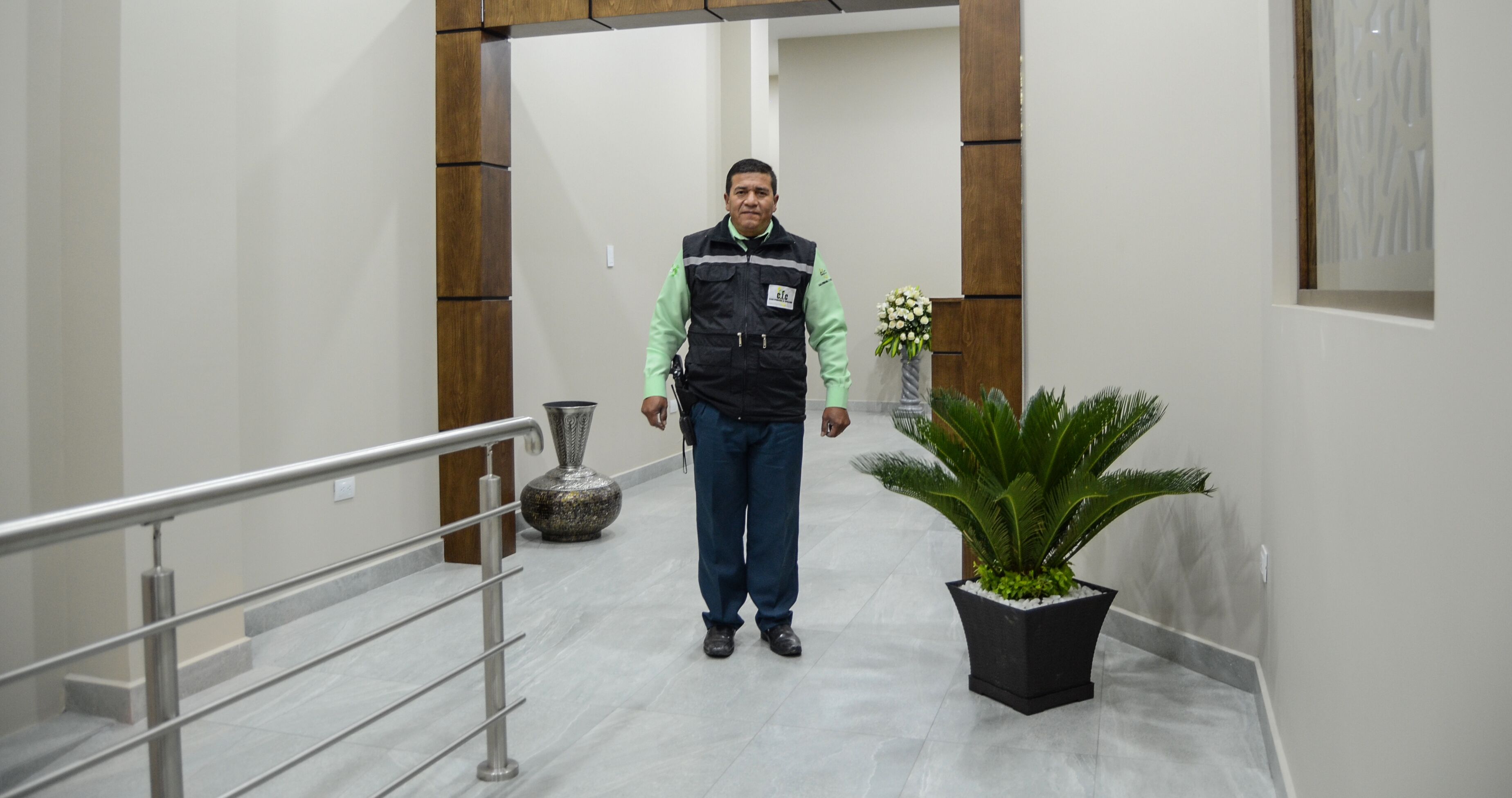 Un hombre está de pie en un pasillo con barandilla metálica, una planta en maceta y un jarrón decorativo.