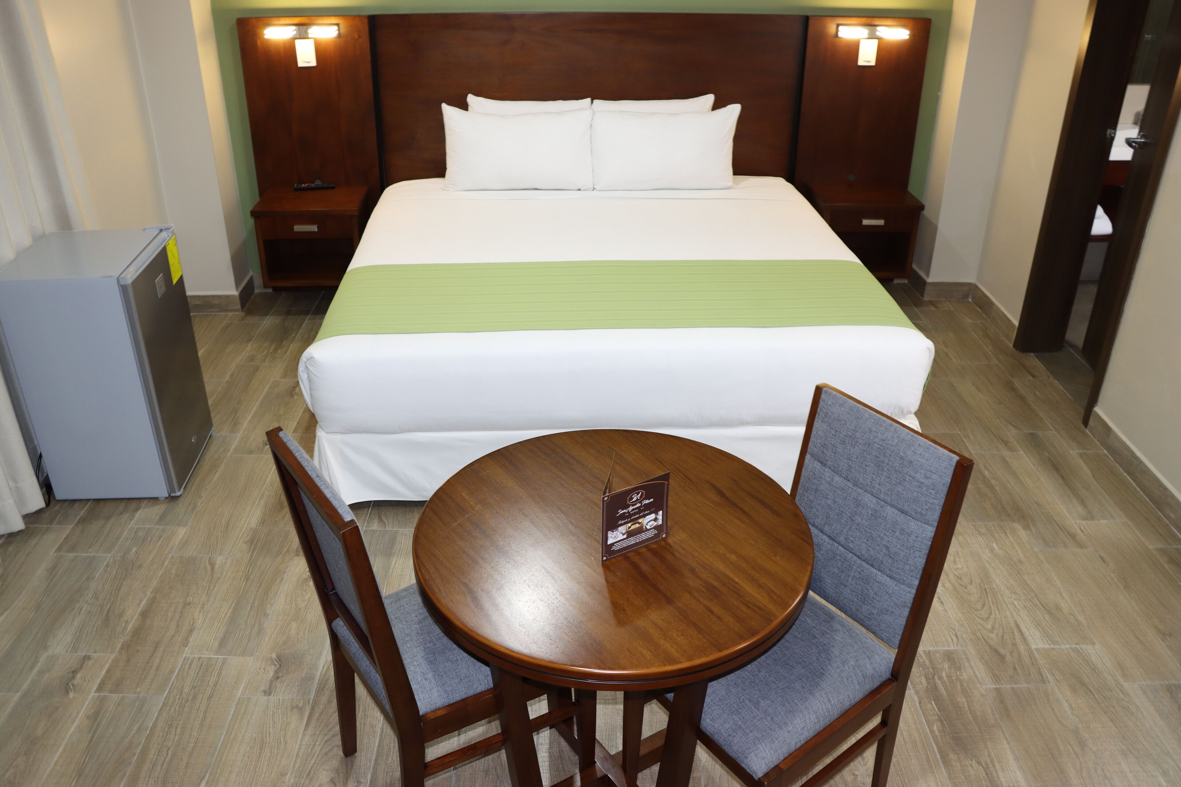 Ein Hotelzimmer mit Bett, einem kleinen Tisch mit Stühlen, einem Kühlschrank und einem Nachttisch mit Lampe.