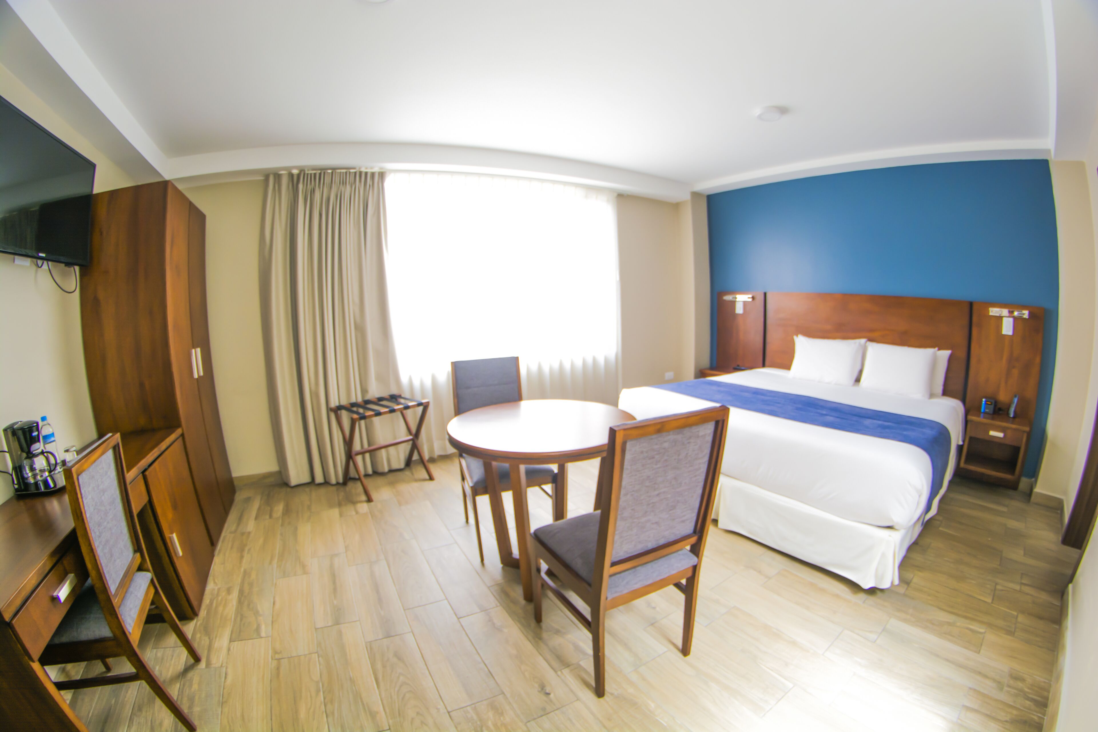 A hotel room with a bed, a desk with a chair, a small table with two chairs, a TV mounted on the wall, and a window with curtains.
