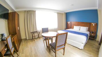 A hotel room with a bed, a desk with a chair, a small table with two chairs, a TV mounted on the wall, and a window with curtains.