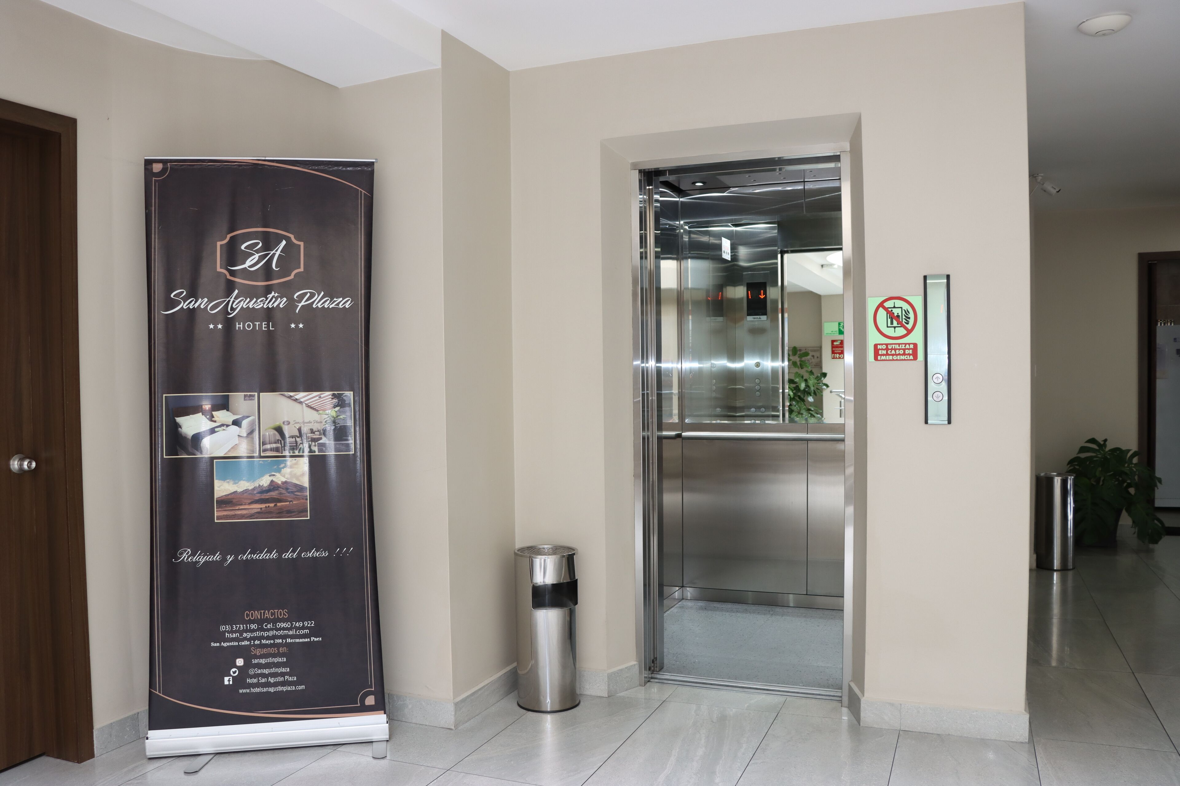 Zona de recepción de hotel con mostrador, un cartel promocional grande, un ascensor y un bote de basura.