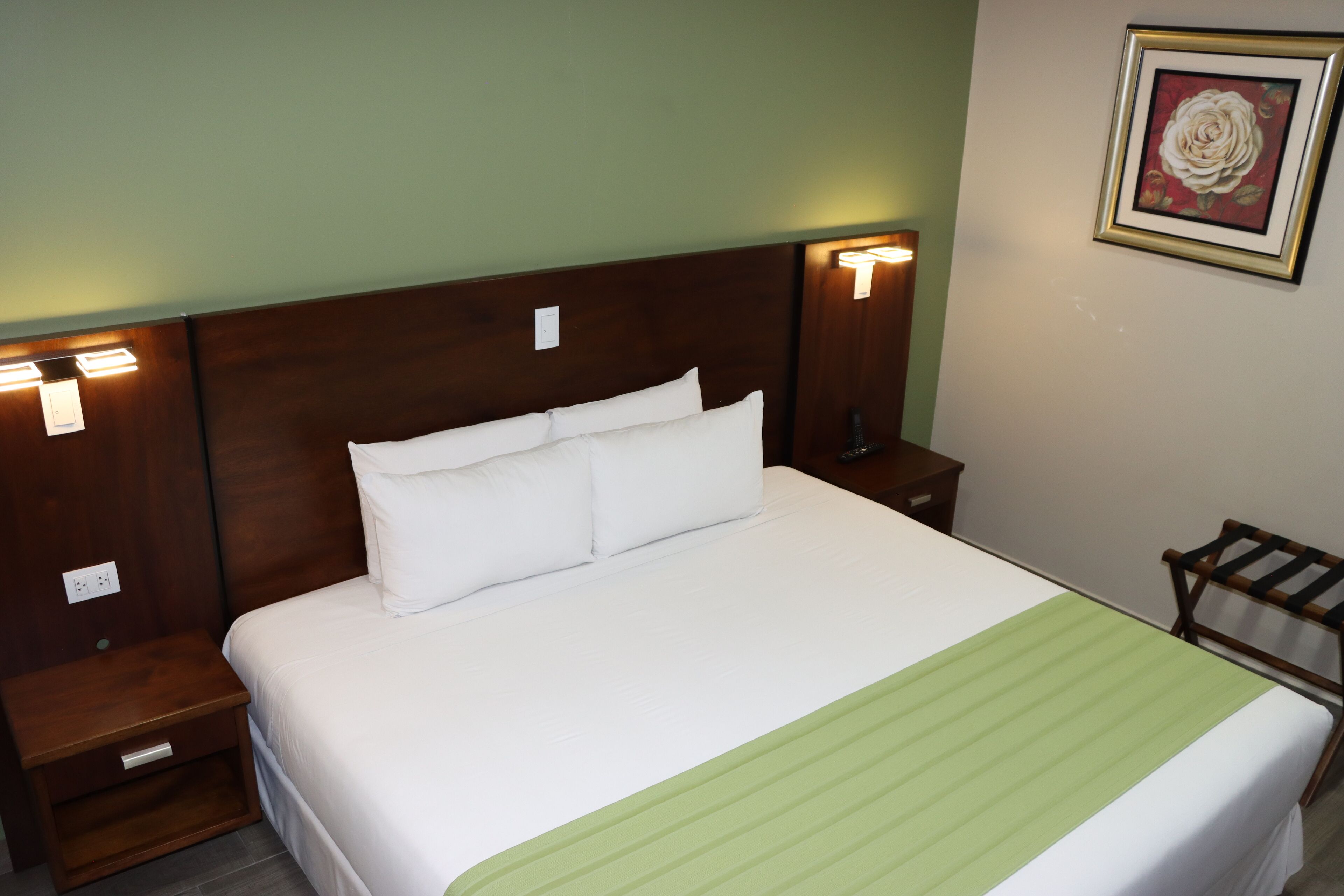 Una cama bien tendida con sábanas blancas y una colcha a rayas verdes, flanqueada por mesitas de noche de madera con lámparas fijadas a la pared.
