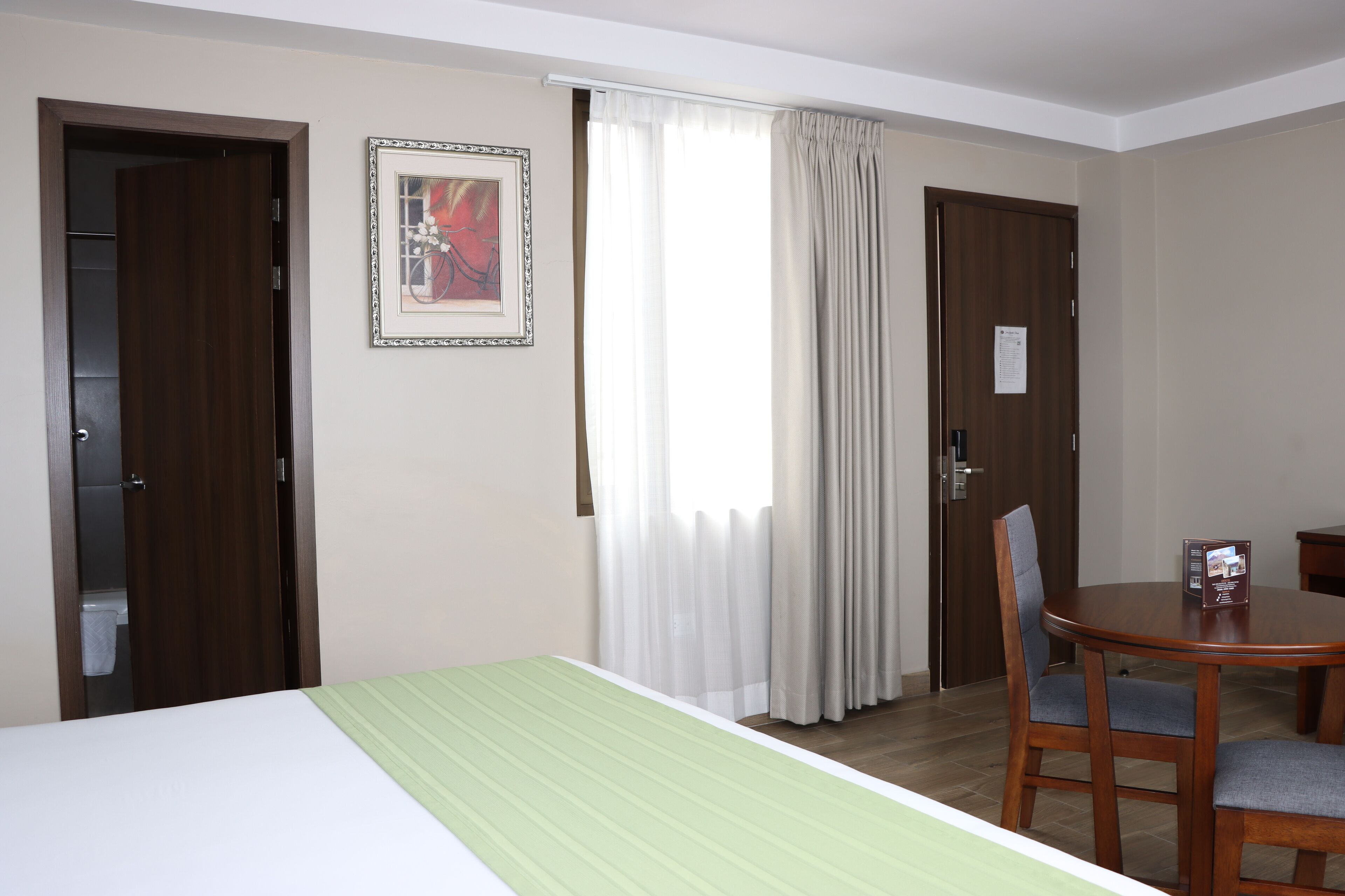 A hotel room with a bed, a dining table with chairs, a window with curtains, and a door.