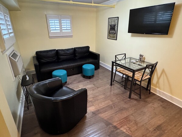 A living room with a black leather sofa, two blue ottomans, a glass dining table with chairs, a mounted flat-screen TV, and a window with blinds. - The Sherman Inn (Batesville)
