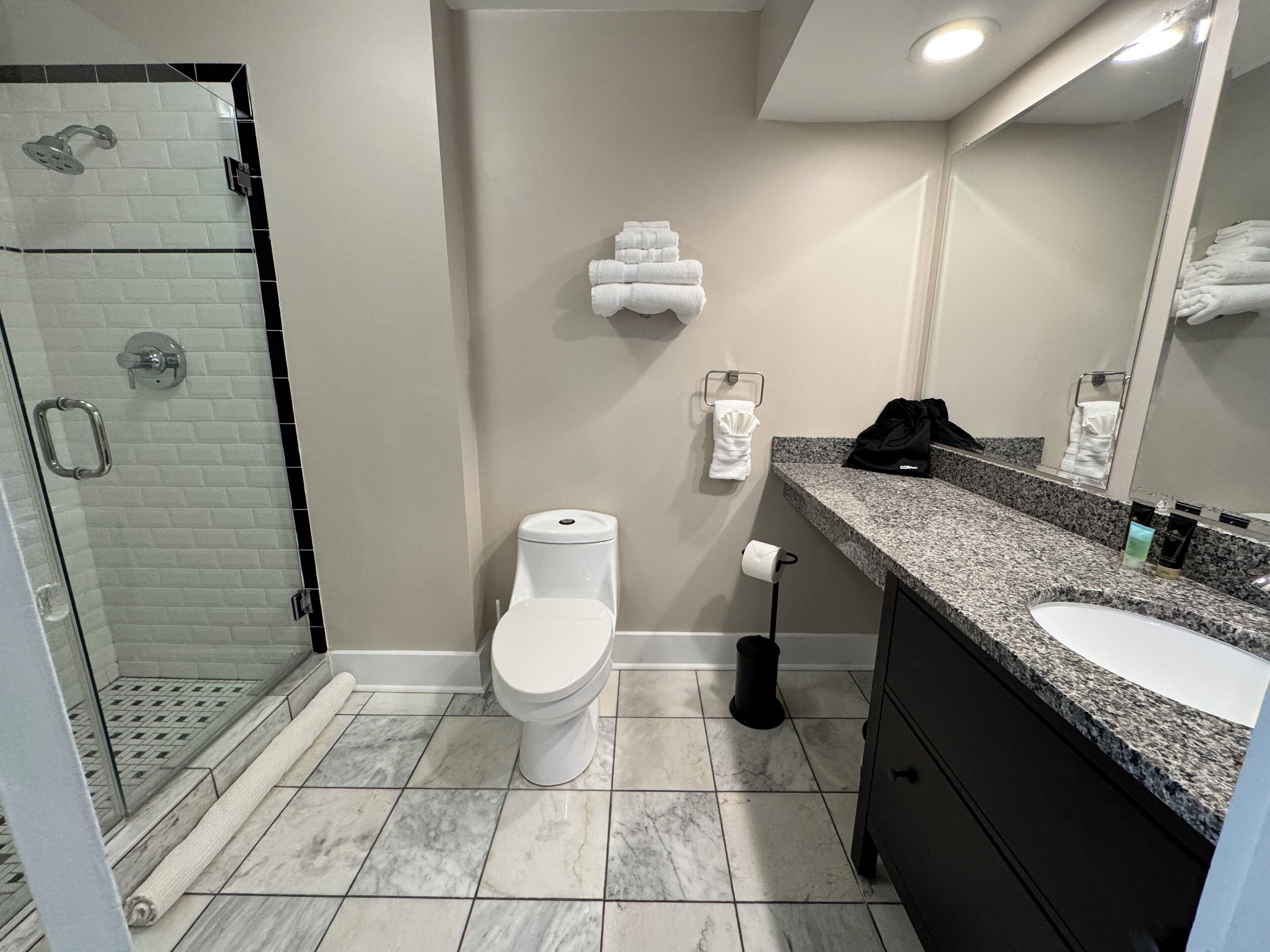 Un baño moderno con cabina de ducha de vidrio, inodoro blanco, suelo de baldosas de mármol, encimera de granito y mueble negro.