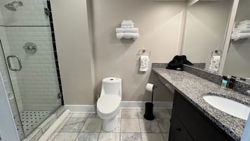 Un baño moderno con cabina de ducha de vidrio, inodoro blanco, suelo de baldosas de mármol, encimera de granito y mueble negro.