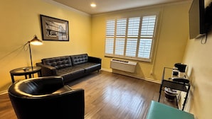 A living room with a black leather sofa, a round coffee table, a floor lamp, a framed picture on the wall, and a window with blinds.