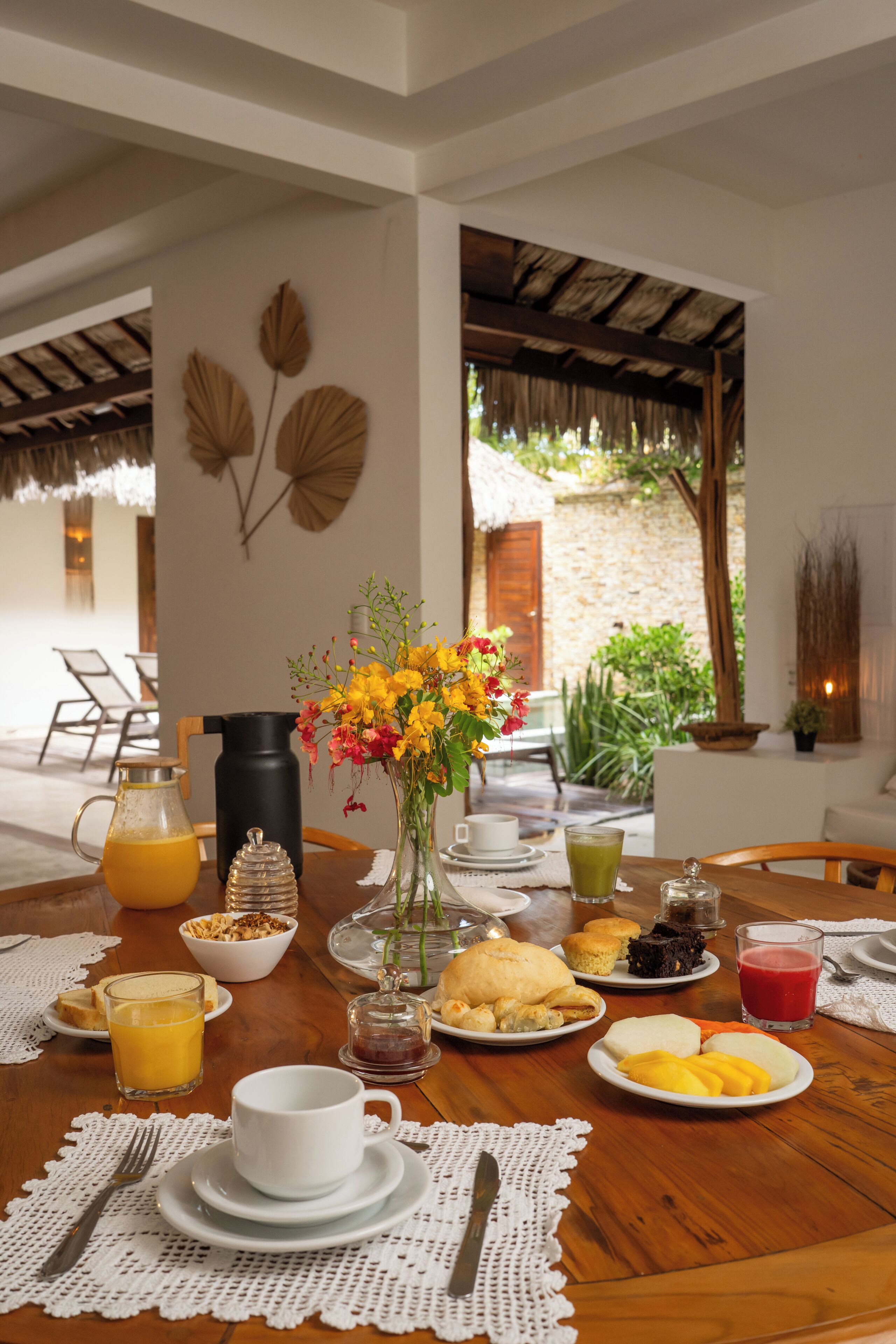 Une table de petit-déjeuner dressée avec divers aliments et boissons, un vase de fleurs et une vue sur un jardin.