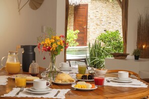 A breakfast table with various food items, a vase of flowers, and a view of a stone wall and greenery outside. - Villa Alegria - La Villa Group (Jijoca de Jericoacoara)