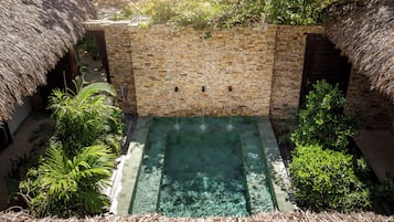 Une piscine à débordement aux murs de pierre, avec une eau cristalline, entourée d’une végétation luxuriante et de constructions aux toits de chaume.