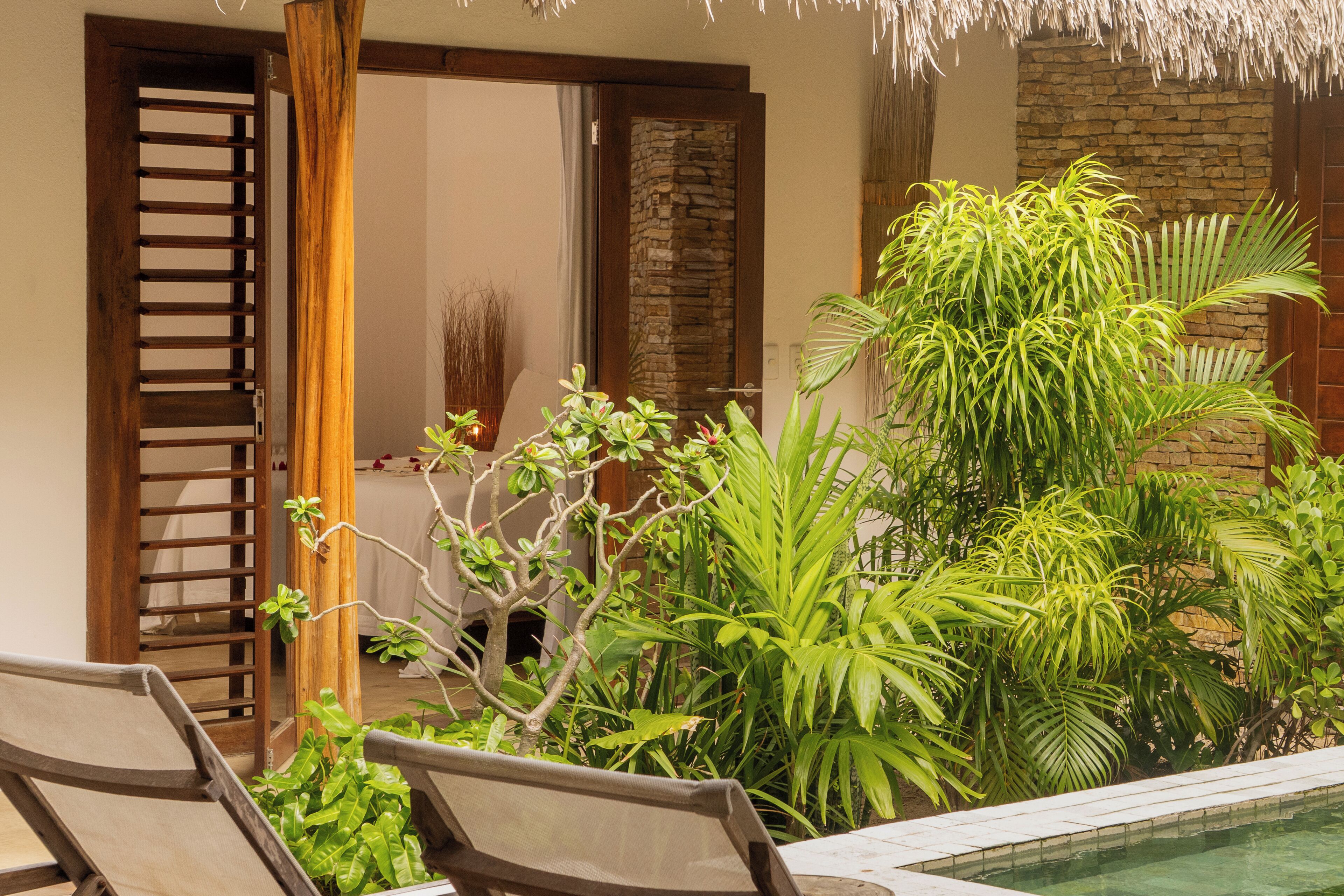 Un área junto a la piscina con sillones de descanso, pared de piedra y plantas tropicales.