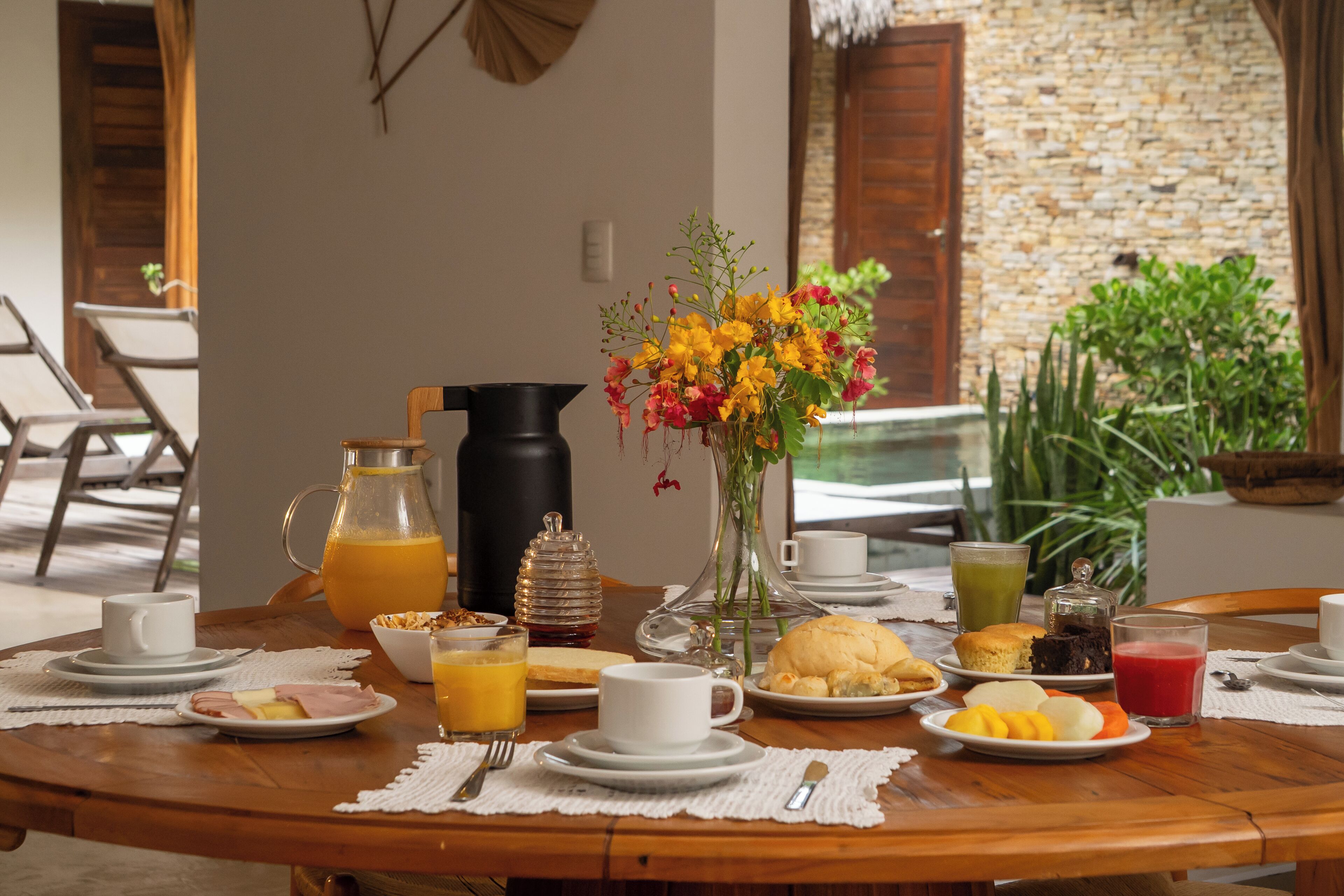 Une table de petit-déjeuner avec divers aliments, un pichet de jus d’orange, un vase rempli de fleurs et une cafetière.