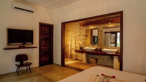 Ein Hotelzimmer mit Bett, Fernseher, einer Holztür, einer Steinwand und einem Badezimmer mit Waschbecken und Spiegel.