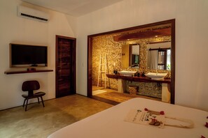 A hotel room with a bed, a television, a wooden door, a stone wall, and a bathroom with a sink and mirror. - Villa Alegria - La Villa Group (Jijoca de Jericoacoara)