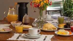A breakfast table with various food items, a pitcher of orange juice, a vase with flowers, and a view of a stone wall and thatched roof in the background. - Villa Alegria - La Villa Group (Jijoca de Jericoacoara)