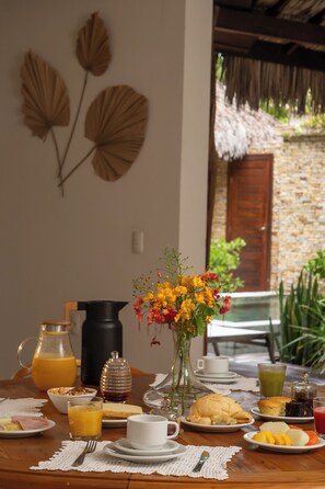 A breakfast table with various food items, a pitcher of orange juice, a vase with flowers, and a view of a stone wall and thatched roof in the background. - Villa Alegria - La Villa Group (Jijoca de Jericoacoara)