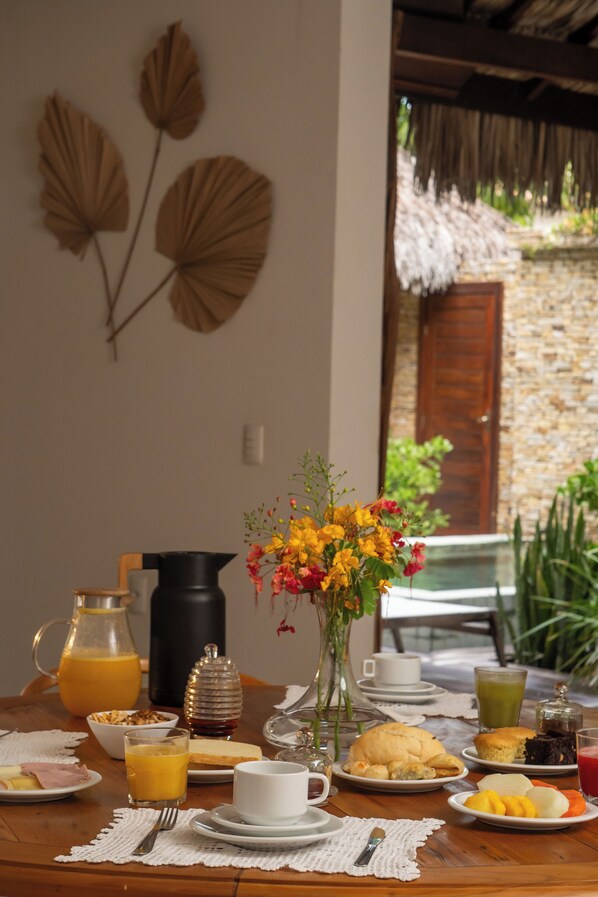 Una mesa de desayuno con diversos alimentos, una jarra de jugo de naranja, un florero con flores y, al fondo, una pared de piedra y un tejado de paja.