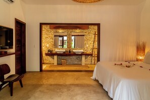 A hotel room with a stone wall, a double sink, a television, and a bed with rose petals. - Villa Alegria - La Villa Group (Jijoca de Jericoacoara)