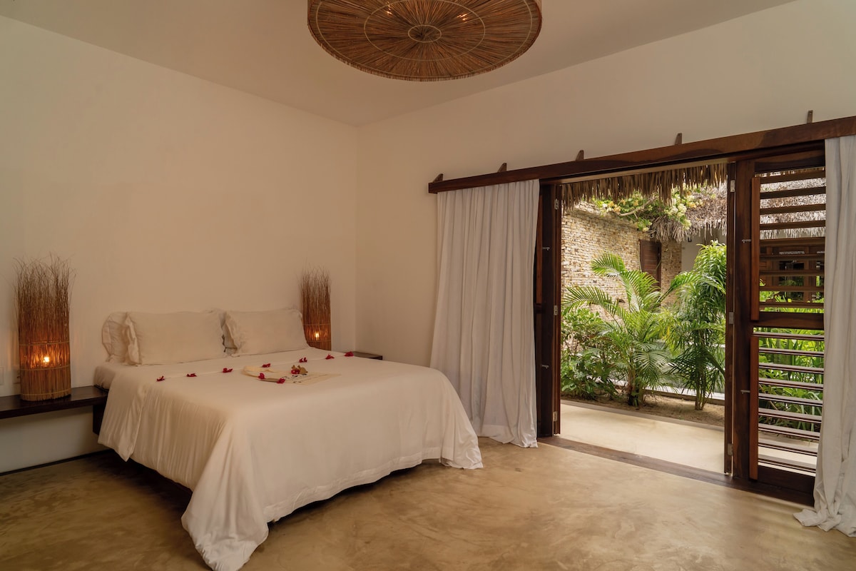 A bedroom with a bed, curtains, a woven ceiling light, and a view of greenery outside.
