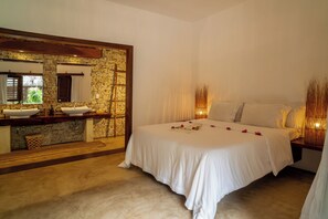 Un dormitorio con pared de piedra, tocador de madera, cama con ropa blanca y un espejo grande.