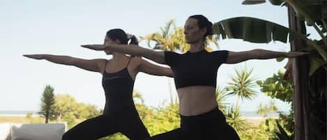 Two individuals performing a yoga pose on a wooden deck with a view of the beach and palm trees in the background.