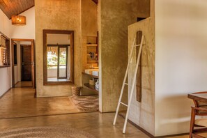 Un pasillo espacioso con suelo de madera, una escalera apoyada en la pared y una puerta que da a una habitación con vistas a la vegetación.