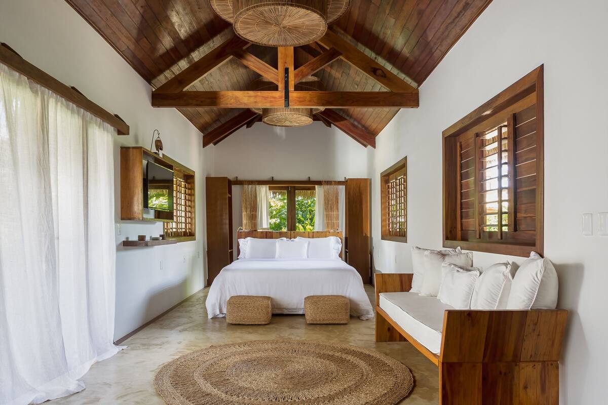 A bedroom with a large bed, wooden ceiling beams, a wooden bench, and a round woven rug.
