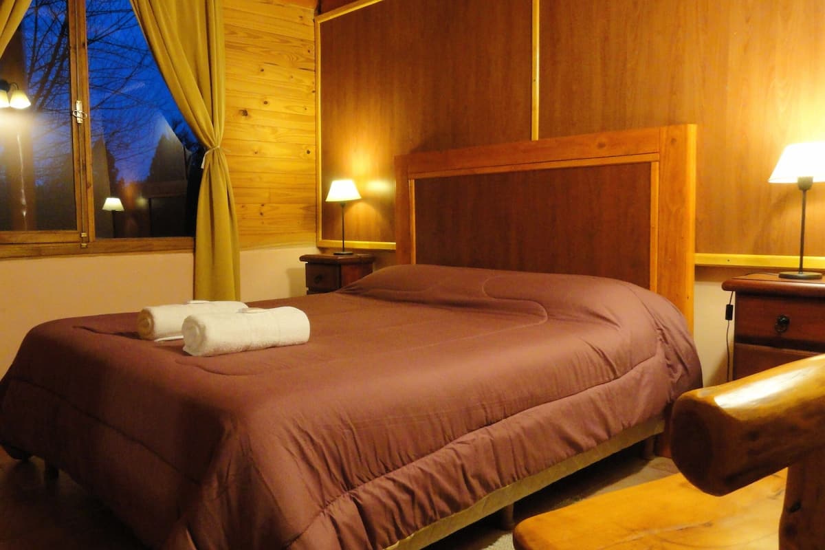 Quarto de madeira com uma cama, duas luminárias de cabeceira e uma janela com cortinas.