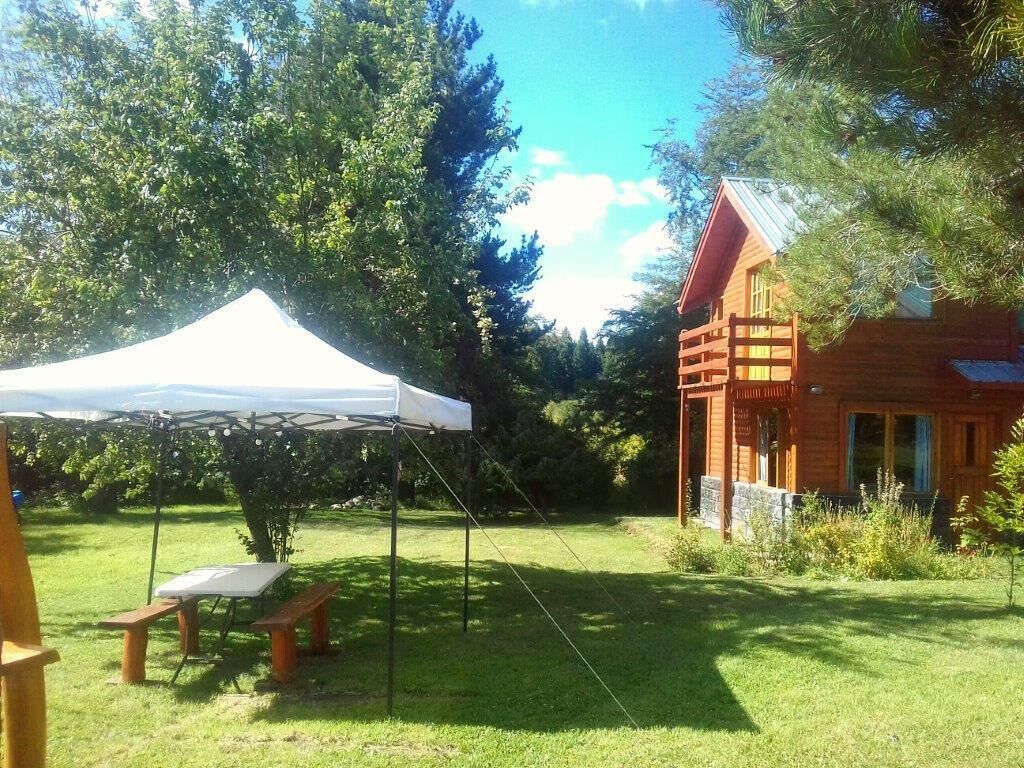 A wooden cabin with a red roof, a white tent, and a picnic table in a grassy area with trees.