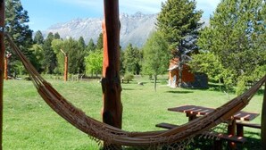 A hammock in a grassy area with a mountain view, picnic tables, and a stone building. - Cabañas El Refugio de Puelo (Lago Puelo)