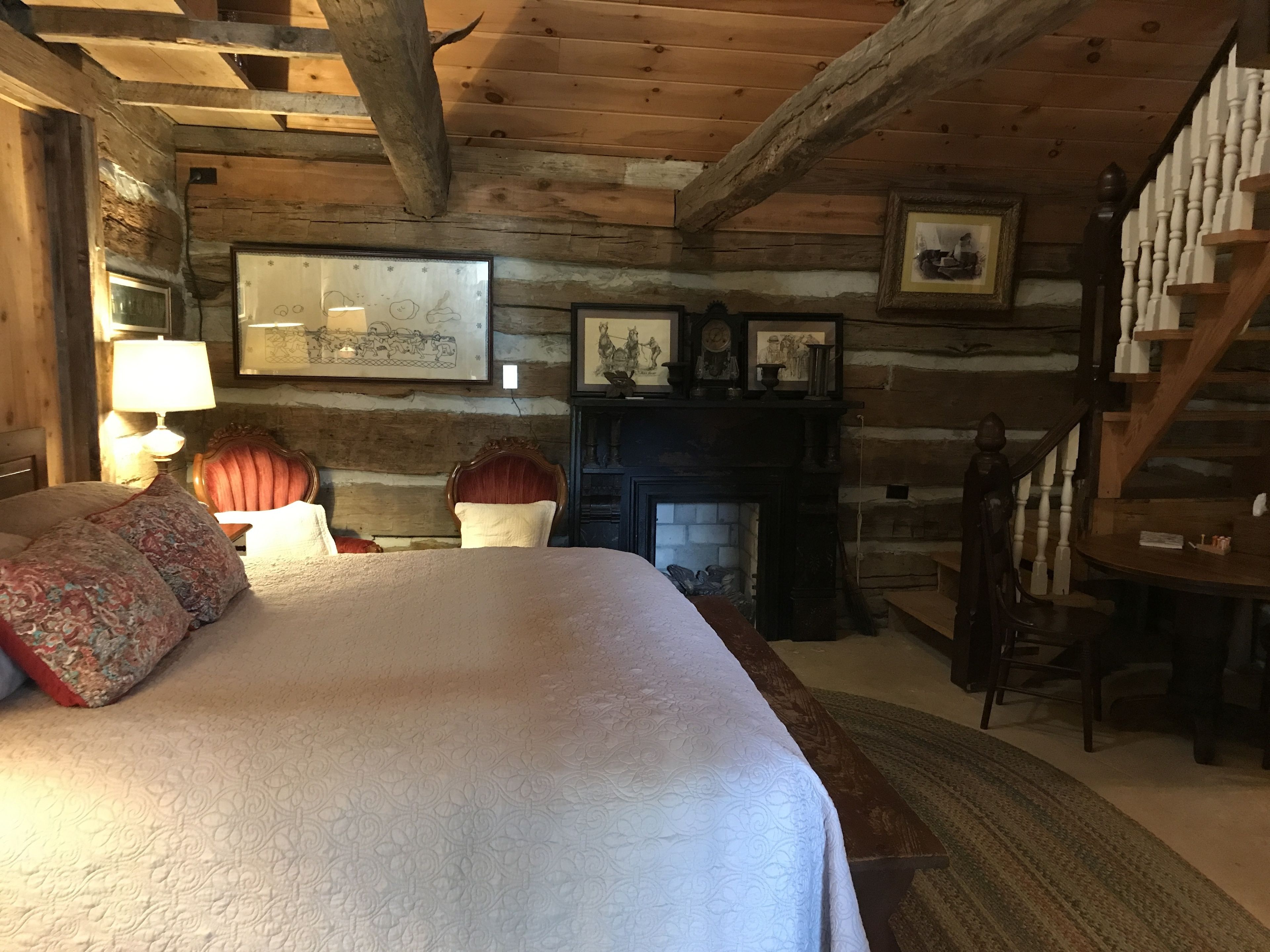 A log cabin bedroom with a large bed, a fireplace, wooden walls, and a staircase.