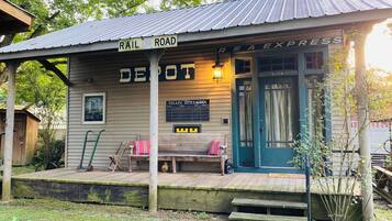 Une gare de train rustique, dotée d’un porche en bois, d’une enseigne indiquant « RAIL ROAD » et d’un panneau d’affichage intitulé « TRAIN BULLETIN ».