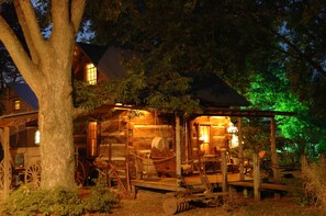 A rustic log cabin with a porch, wooden furniture, and a large tree in the foreground. - Pecan Grove Cottages (Martin)