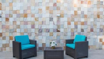 Duas cadeiras de vime com almofadas turquesa e uma pequena mesa com um vaso de flores em frente a uma parede de mosaico.