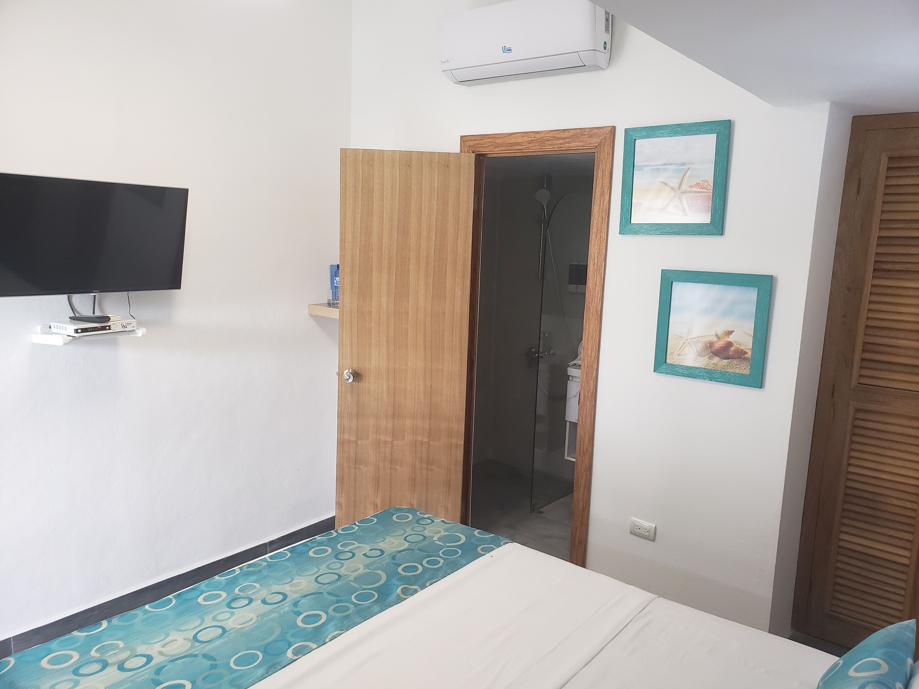 Quarto com cama, televisão na parede, ar condicionado e porta de madeira para o banheiro.