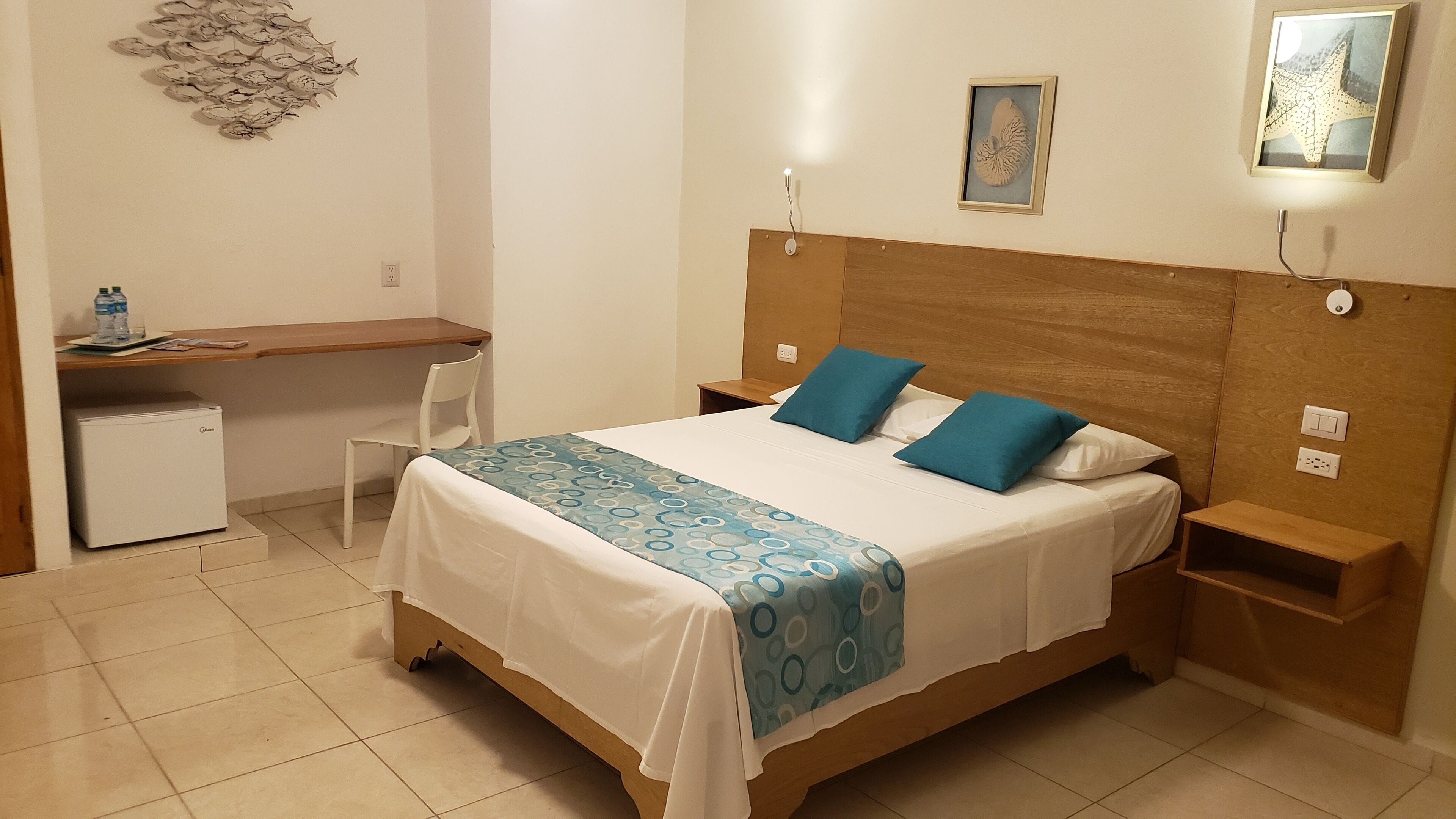 Un dormitorio con cama, escritorio, silla, refrigerador pequeño e iluminación de pared.