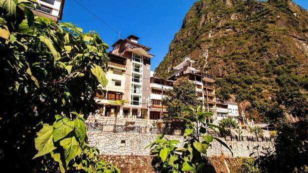 Un hotel de varios pisos con balcones, un muro de piedra de contención y montañas al fondo.