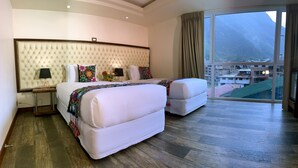 Habitación de hotel con una cama grande, una cama más pequeña, una mesita de noche, una lámpara y vistas a un paisaje montañoso a través de un ventanal amplio.