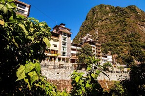 Um hotel com vários andares, varandas, um muro de pedra e montanhas ao fundo.