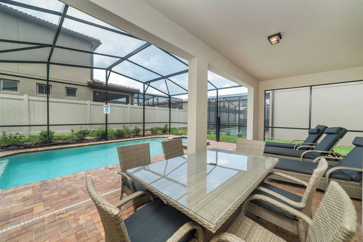 A covered patio with a glass table and wicker chairs, a pool, and a white fence.