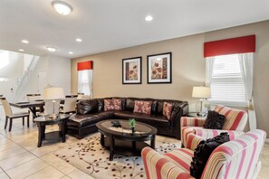 A living room with a sectional sofa, a round coffee table, a striped armchair, and a dining area with a table and chairs. - Windsor at Westside Resort Homes (Kissimmee)