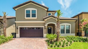 A two-story house with a brown garage door, a front entrance, and a landscaped garden with palm trees. - Windsor at Westside Resort Homes (Kissimmee)